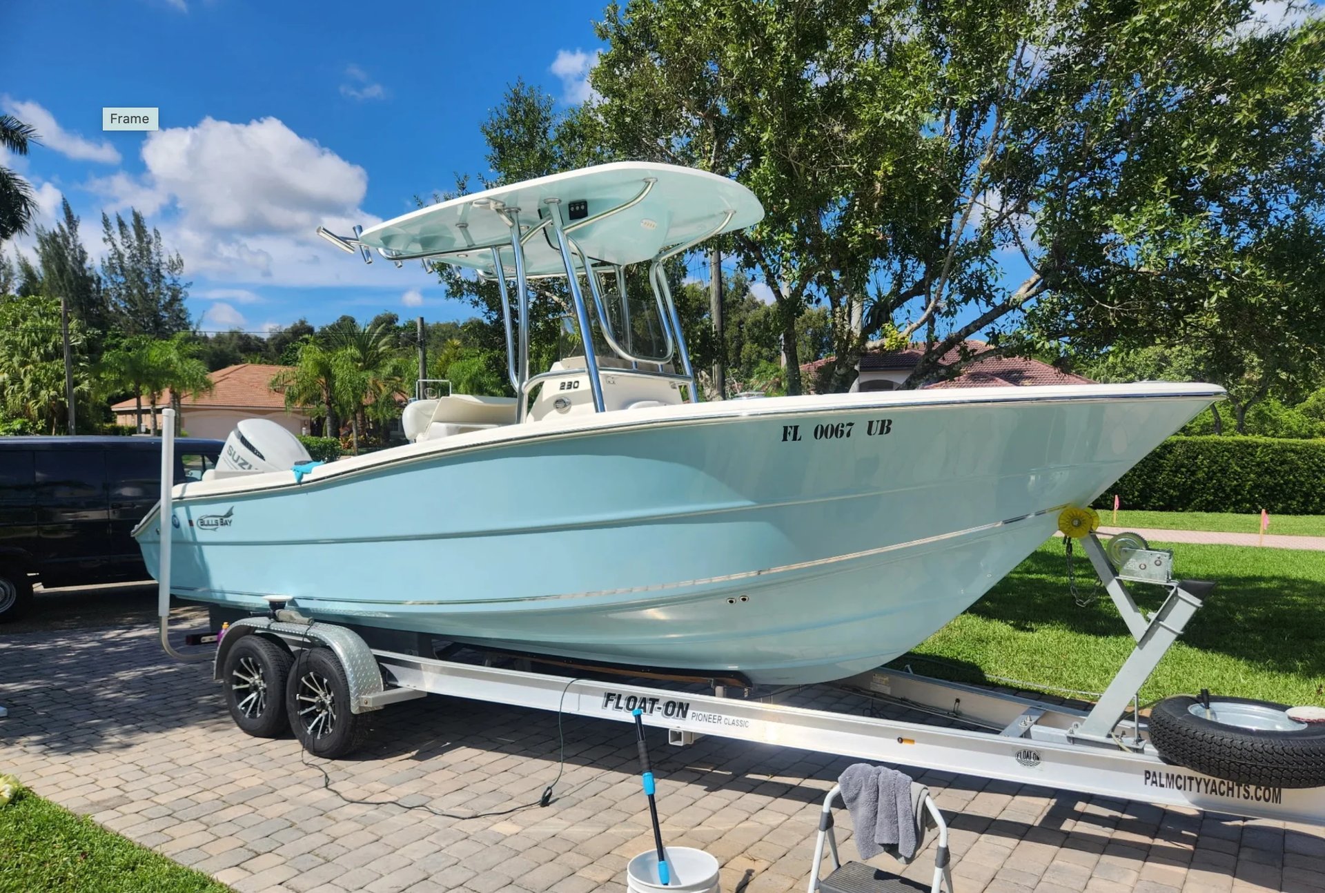 Light blue and white fishing boat on a dual-axle trailer parked in a residential driveway with grass and trees in background